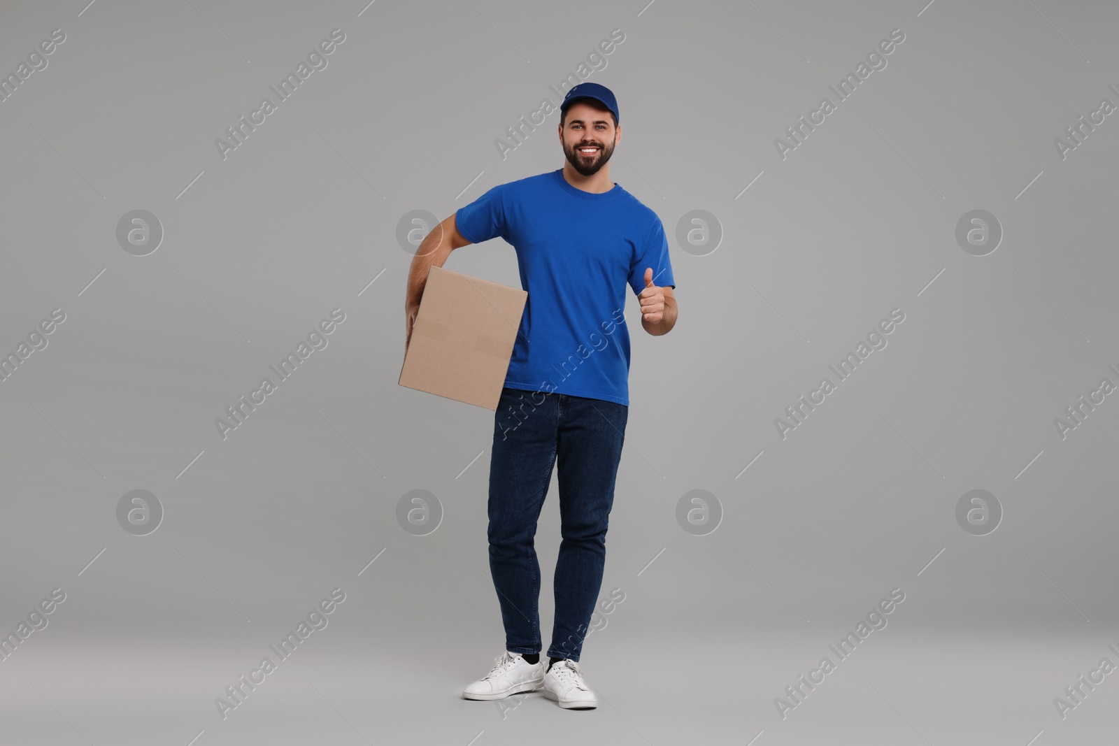 Photo of Happy young courier with parcel showing thumb up on grey background