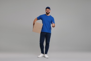 Happy young courier with parcel showing thumb up on grey background