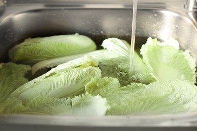 Photo of Pouring tap water on Chinese cabbage leaves in sink, closeup