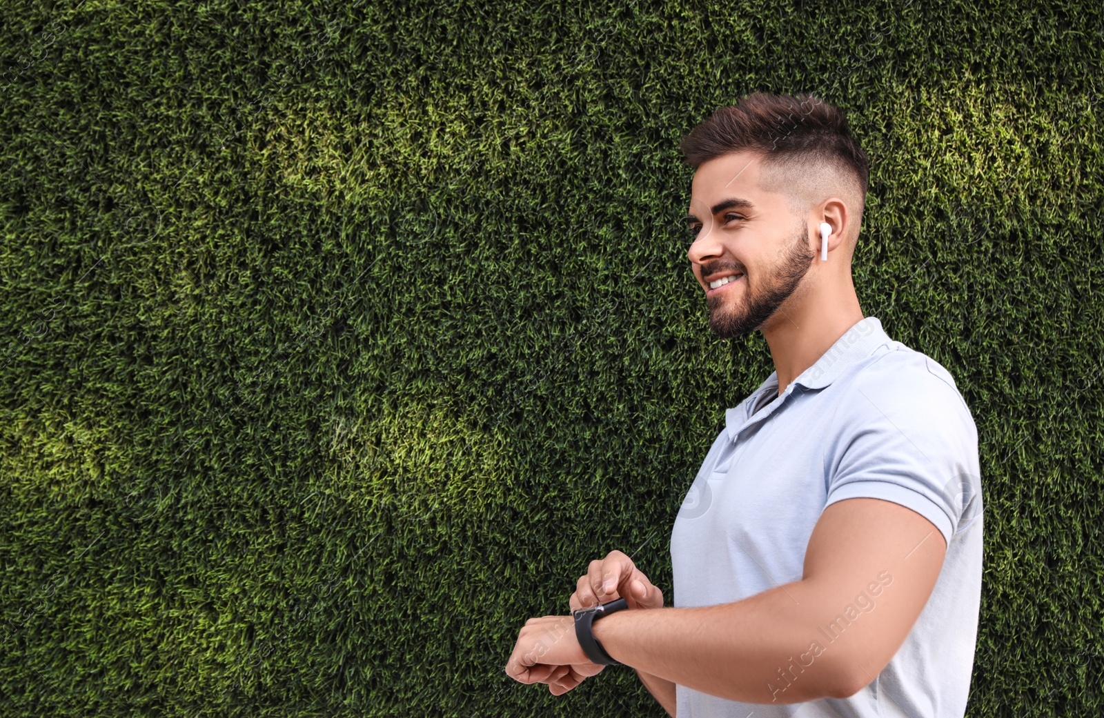 Photo of Young man with wireless headphones and smartwatch listening to music near green grass wall. Space for text