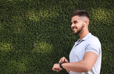 Young man with wireless headphones and smartwatch listening to music near green grass wall. Space for text