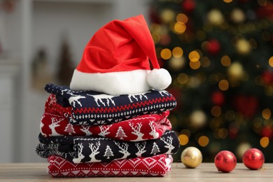 Photo of Stack of different Christmas sweaters, Santa Claus hat and decorative balls on table against blurred lights