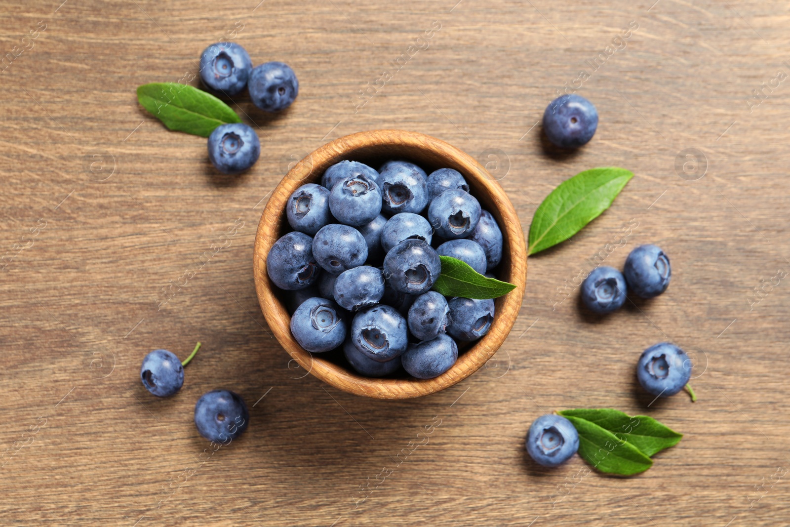 Photo of Bowl of tasty fresh blueberries with green leaves on wooden table, flat lay