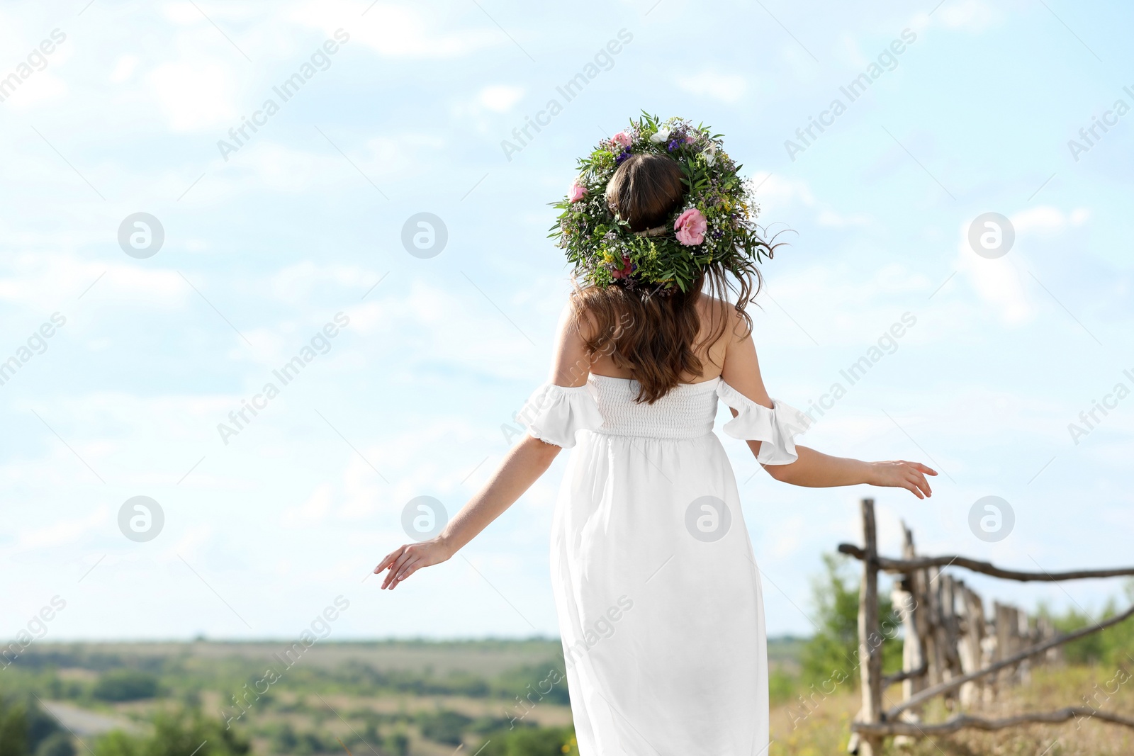 Photo of Young woman wearing wreath made of beautiful flowers outdoors on sunny day, back view