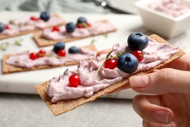 Photo of Woman holding tasty cracker sandwich with cream cheese, blueberries, red currants and thyme at table, closeup
