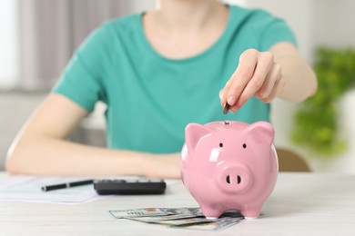 Financial savings. Woman putting coin into piggy bank at white wooden table indoors, closeup