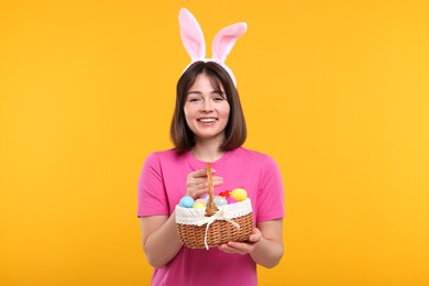 Photo of Easter celebration. Happy woman with bunny ears and wicker basket full of painted eggs on orange background