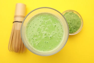 Photo of Glass of tasty matcha smoothie, powder and bamboo whisk on yellow background, flat lay