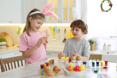 Photo of Easter celebration. Cute children with bunny ears painting eggs at white marble table in kitchen