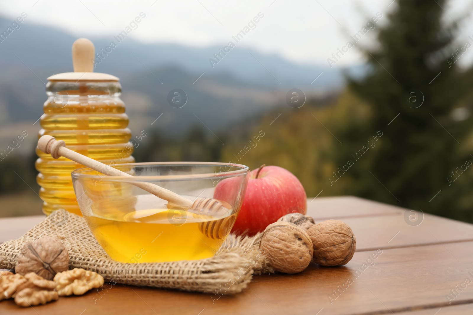 Photo of Fresh aromatic honey, apple and walnuts on wooden table against mountain landscape. Space for text