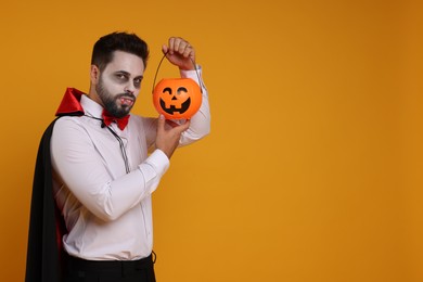 Photo of Man in scary vampire costume with fangs and pumpkin bucket on orange background, space for text. Halloween celebration