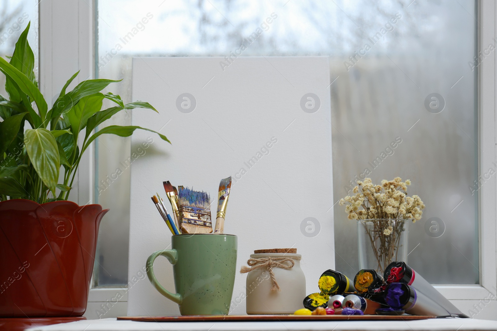 Photo of White canvas, colorful paints and brushes in cup on table near window