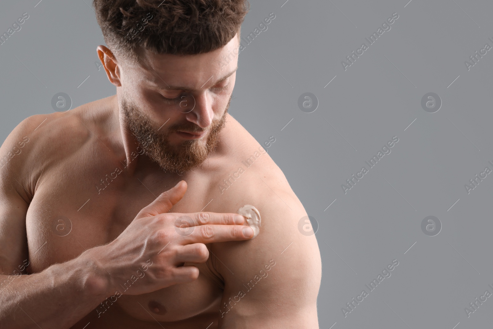 Photo of Handsome man applying moisturizing cream onto his shoulder on grey background, space for text