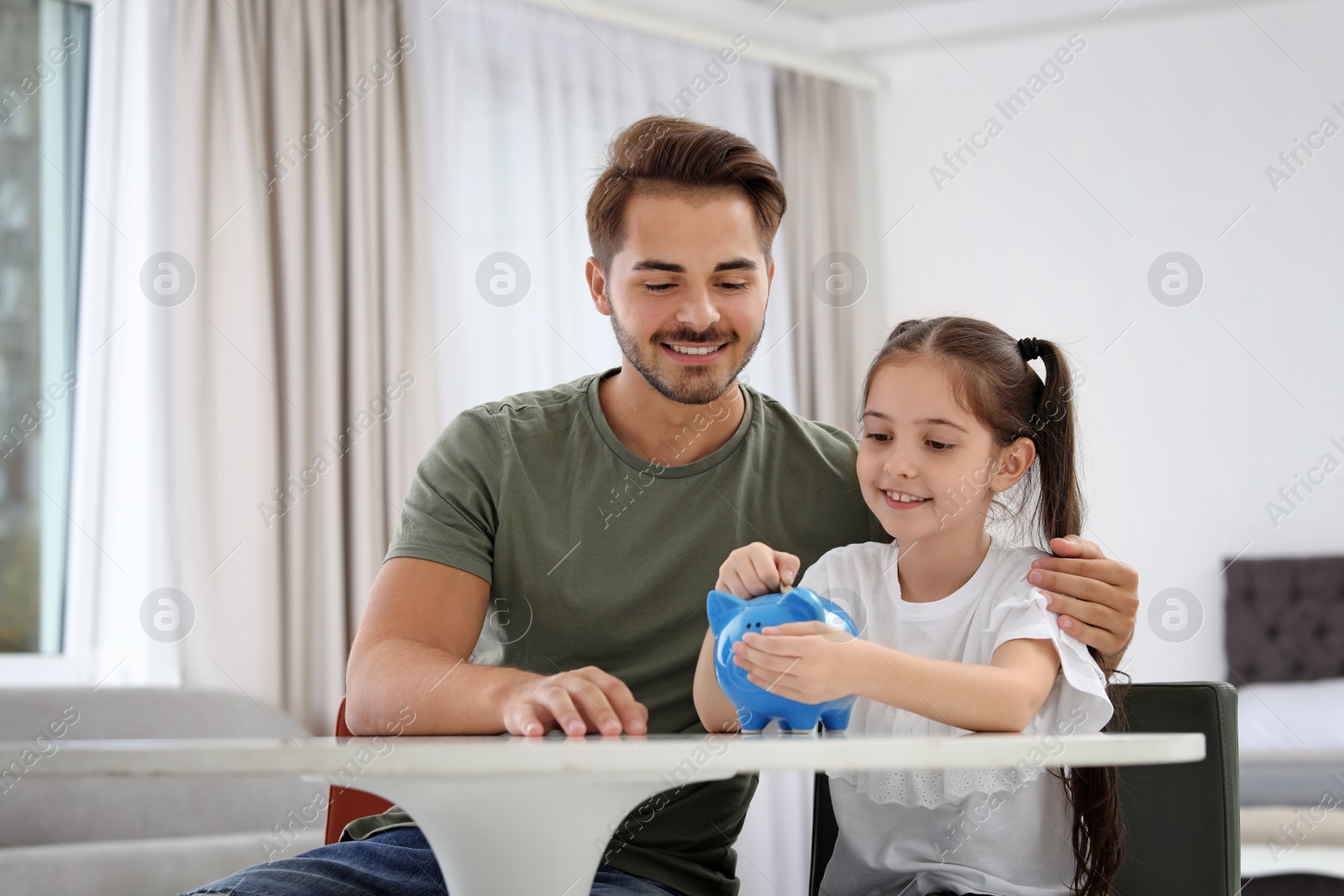 Photo of Little girl with her father putting coin into piggy bank at table indoors