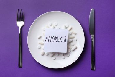 Photo of Cutlery near plate with pills and word Anorexia on dark violet background, flat lay