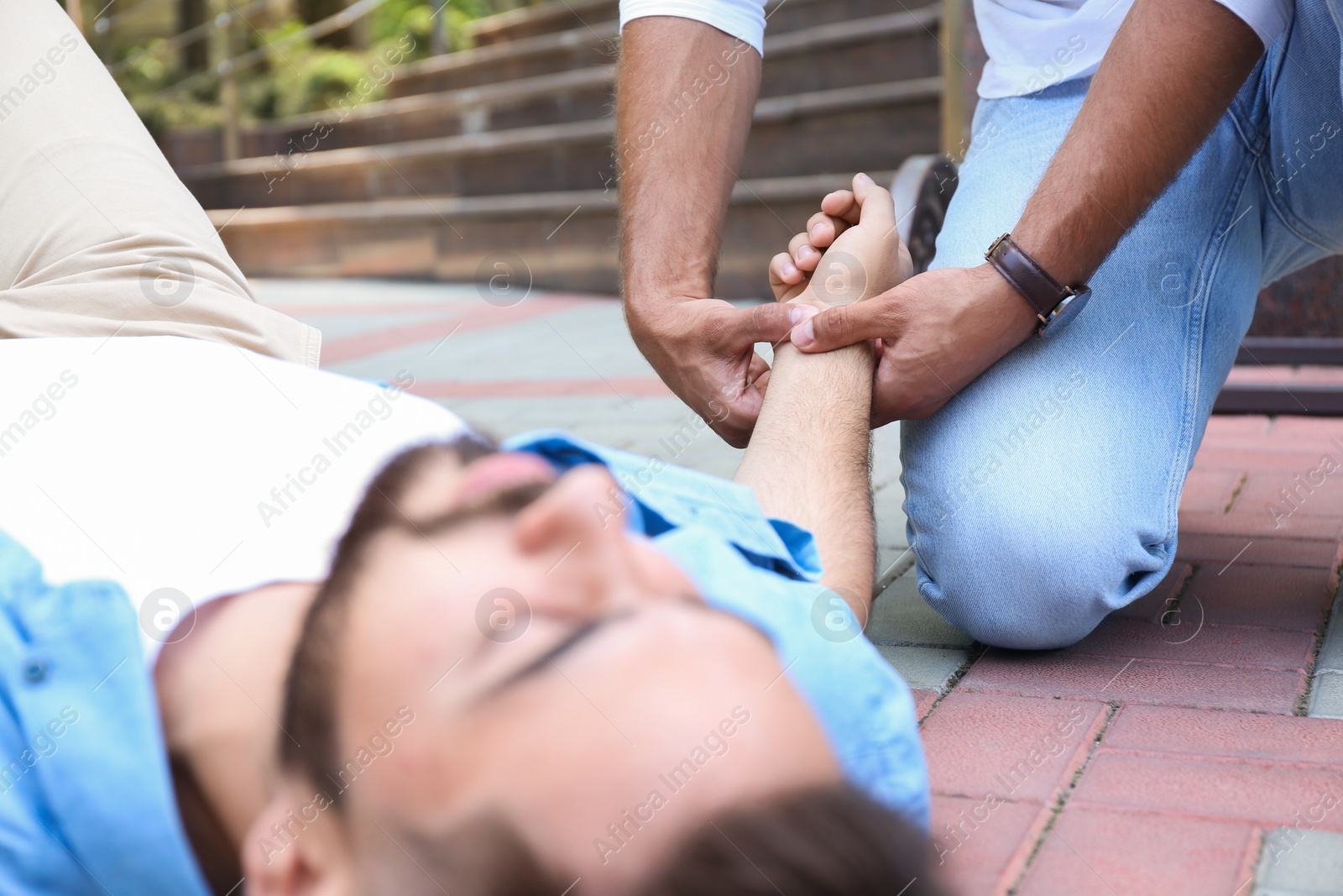Photo of Passerby checking pulse of unconscious young man outdoors, closeup. First aid
