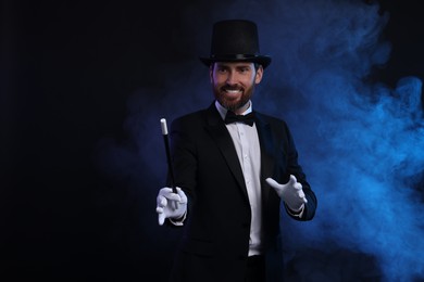 Photo of Happy magician holding wand in smoke on dark background