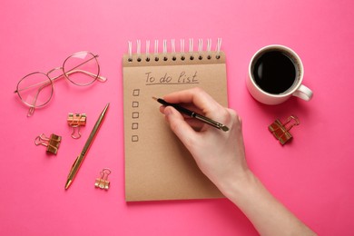Photo of Woman filling To Do list in notepad at pink table, top view