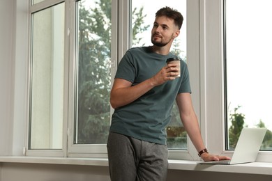 Photo of Handsome man with laptop and cup of coffee near window indoors, space for text