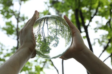 Photo of Overturned reflection of beautiful green trees outdoors, low angle view. Man holding crystal ball in park, closeup