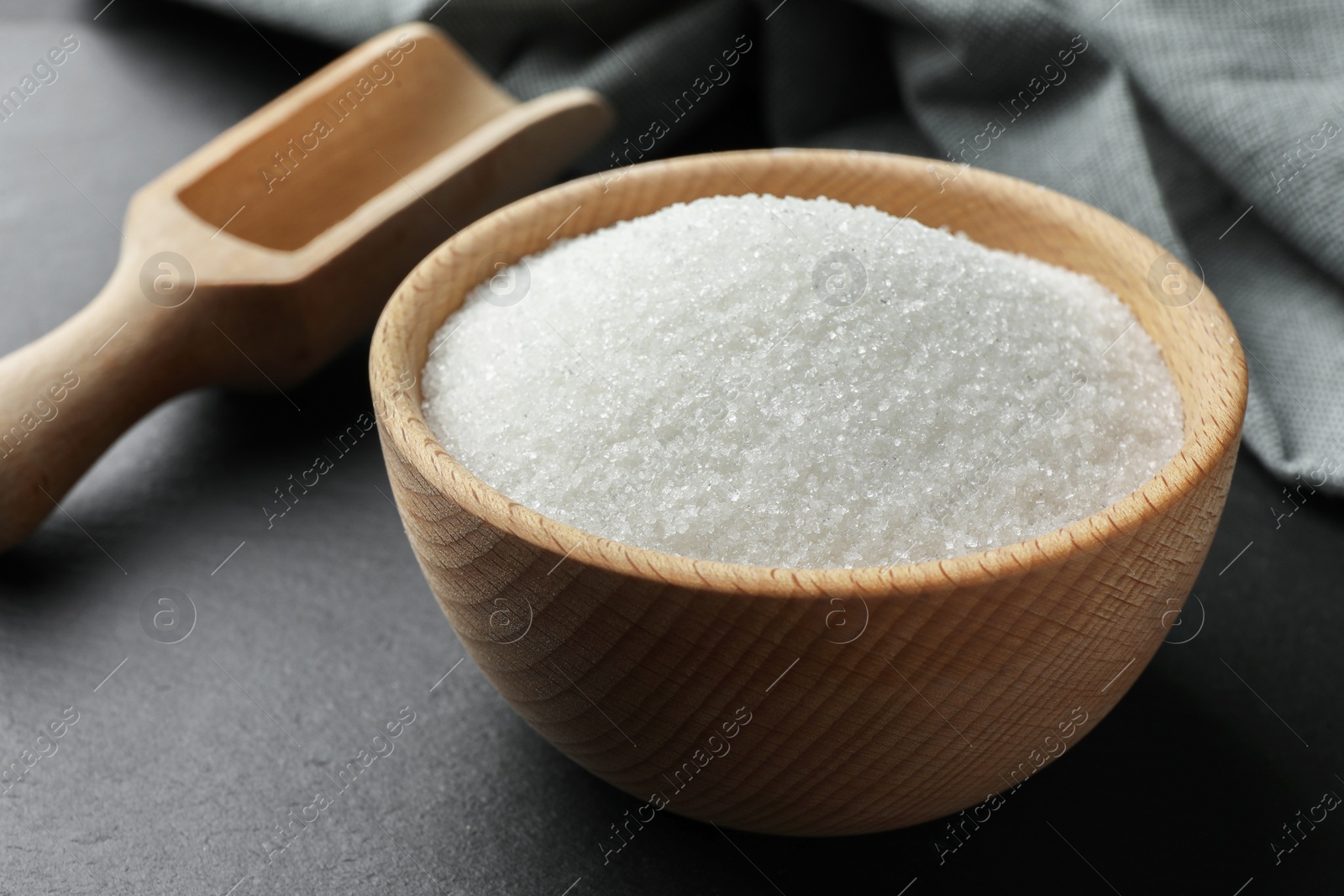 Photo of Granulated sugar in bowl and wooden scoop on black table, closeup