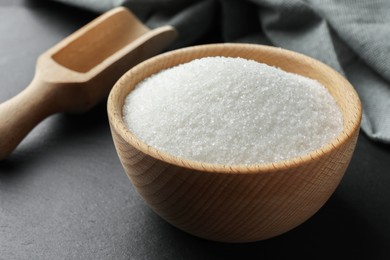 Granulated sugar in bowl and wooden scoop on black table, closeup