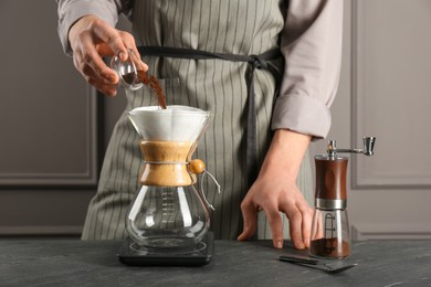 Photo of Man adding coffee into glass chemex coffeemaker with paper filter at gray table, closeup