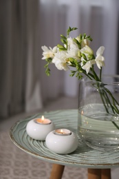 Photo of Vase with beautiful freesia flowers and burning candles on stand indoors, closeup. Interior elements