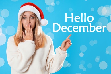Image of Hello December greeting card. Surprised woman in Santa hat on light blue background, bokeh effect
