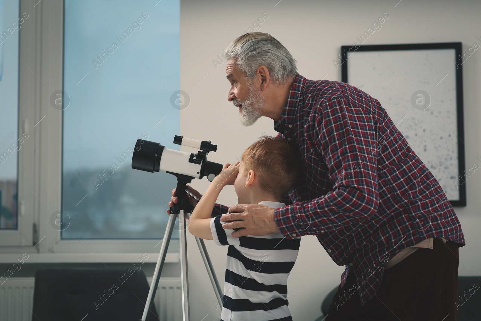 Photo of Little boy with his grandfather looking at stars through telescope in room