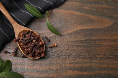 Spoon with aromatic cloves and green leaves on wooden table, flat lay. Space for text