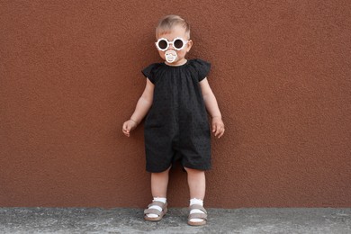 Photo of Cute little girl wearing stylish clothes with sunglasses and pacifier near brown wall outdoors