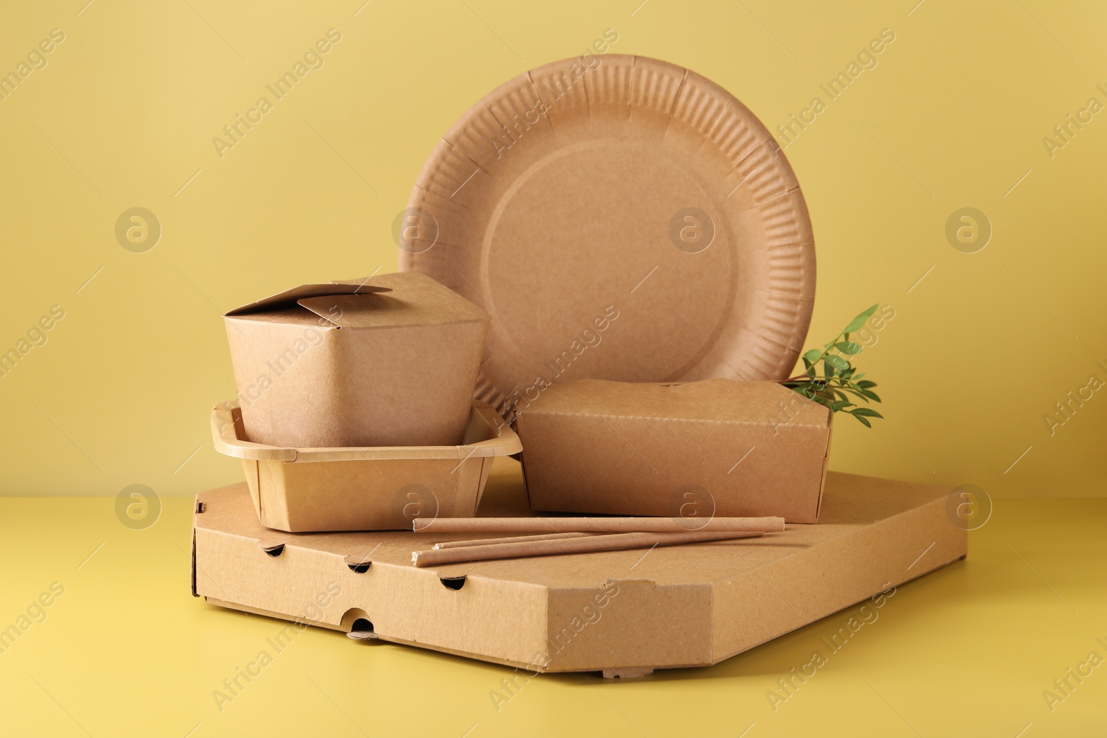 Photo of Eco friendly food packaging. Paper containers, tableware and green twig on pale yellow background