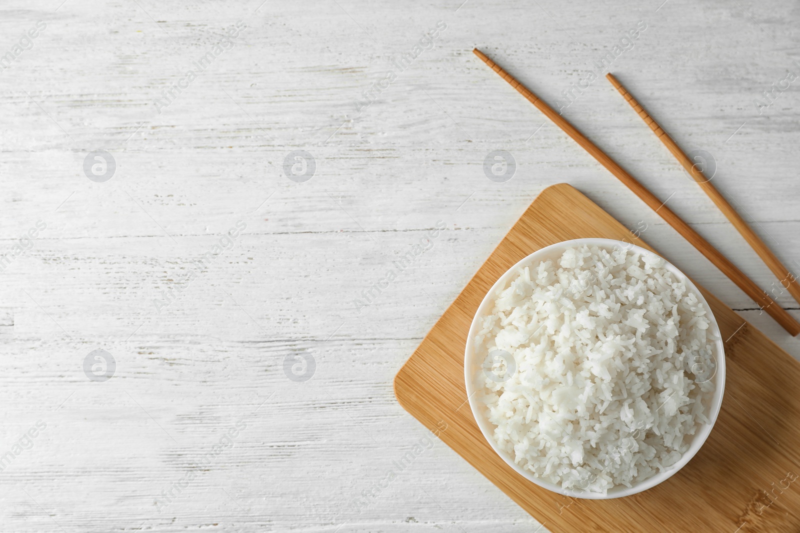 Photo of Board with boiled rice in bowl and chopsticks on wooden background, top view. Space for text