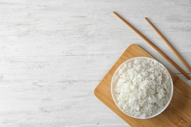 Photo of Board with boiled rice in bowl and chopsticks on wooden background, top view. Space for text