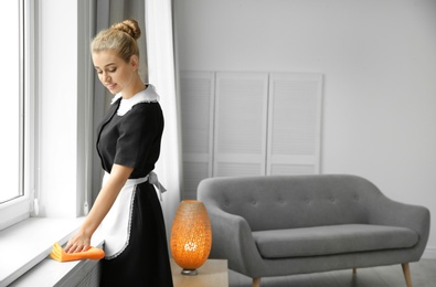 Photo of Young chambermaid cleaning window sill with rag indoors. Space for text
