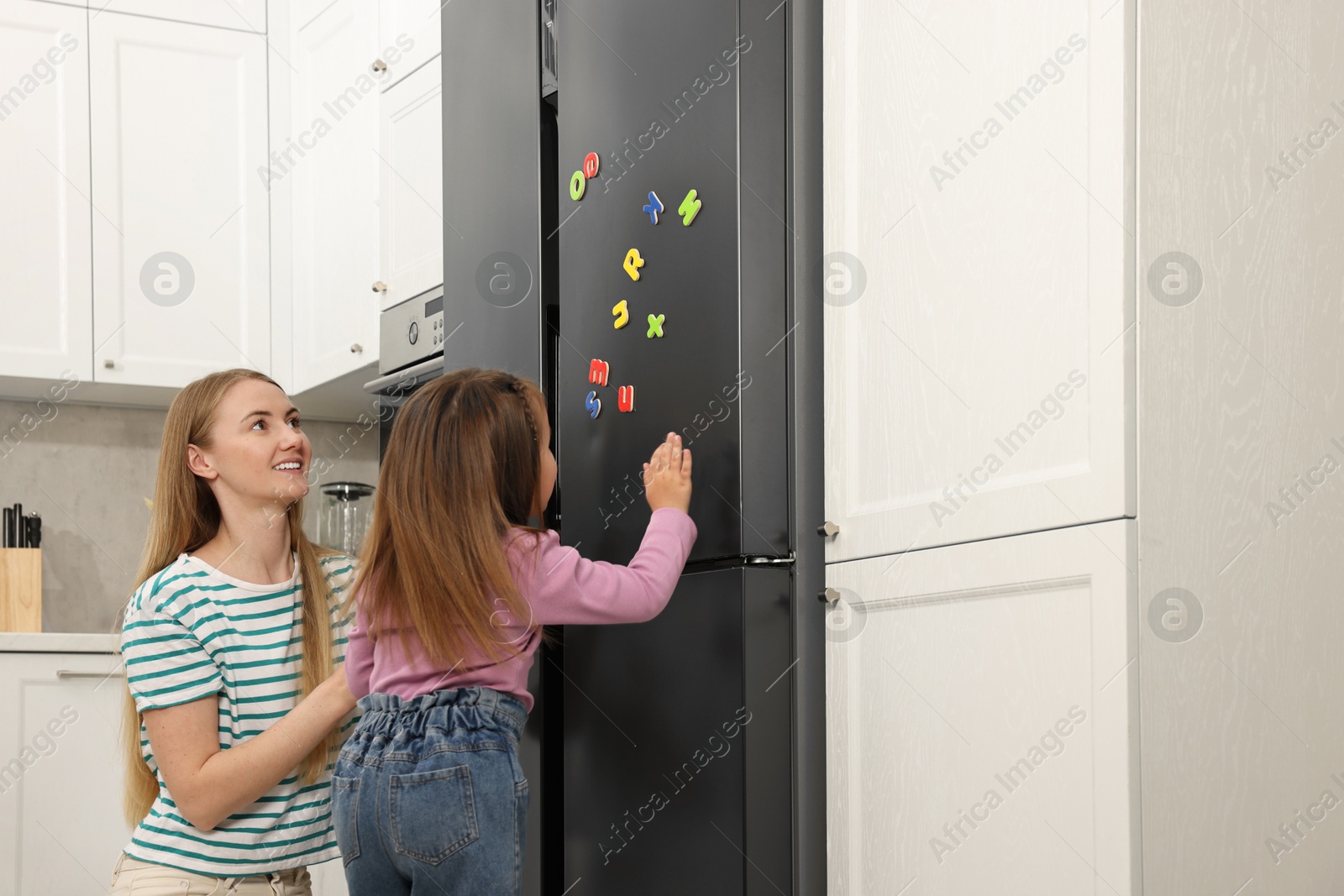 Photo of Mom and daughter putting magnetic letters on fridge at home. Learning alphabet