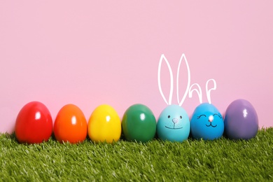 Two eggs with drawn faces and ears as Easter bunnies among others on green grass against pink background