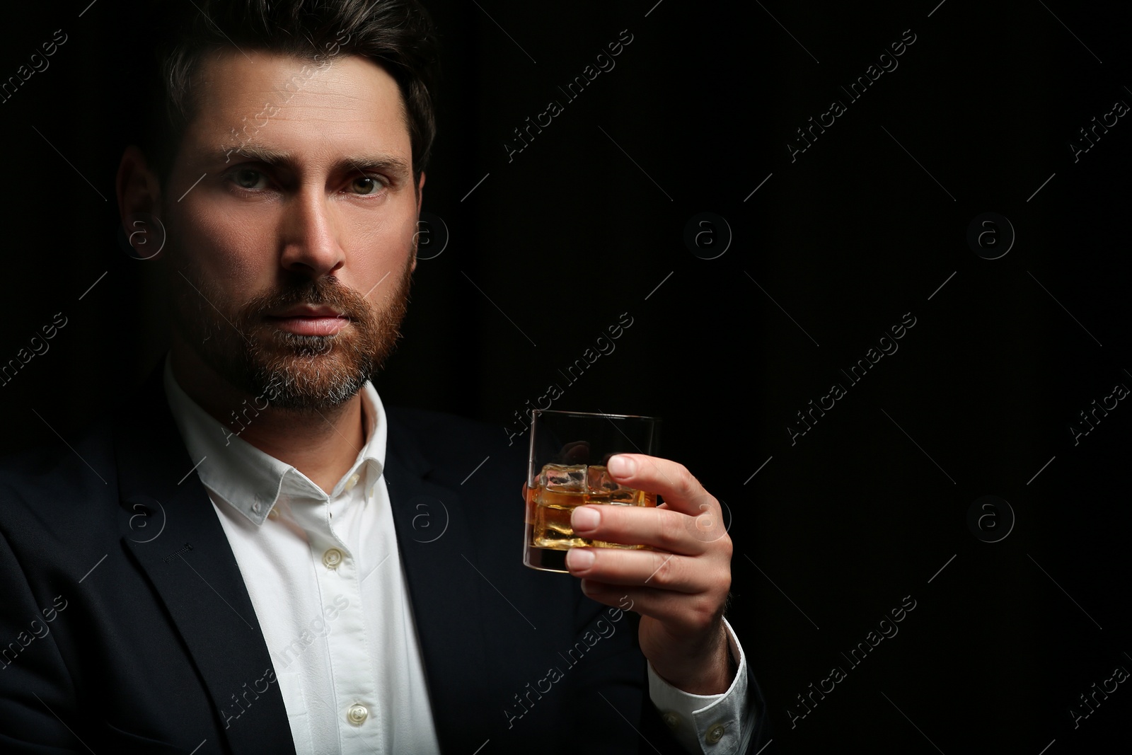 Photo of Man in suit holding glass of whiskey with ice cubes on black background. Space for text