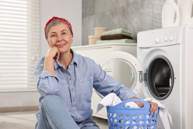 Photo of Happy housewife with laundry basket near washing machine at home