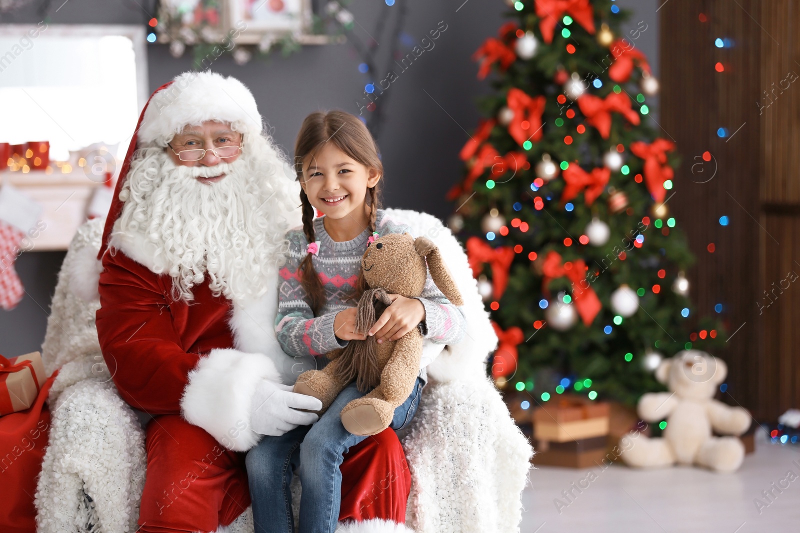 Photo of Little girl with toy bunny sitting on authentic Santa Claus' lap indoors