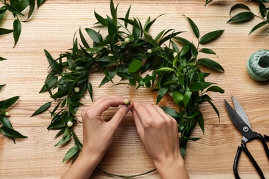 Photo of Florist making beautiful mistletoe wreath at wooden table, top view. Traditional Christmas decor