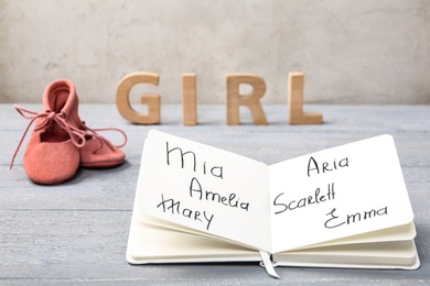 Photo of Notebook with written different baby names, child's shoes and wooden letters on grey wooden table