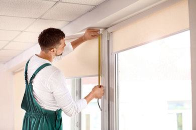 Handyman with tape measure installing roller window blind indoors