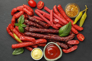 Photo of Different thin dry smoked sausages, basil and sauces on black table, flat lay