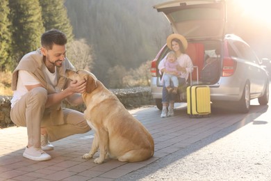 Photo of Happy man petting dog, mother and her daughter sitting in car trunk outdoors. Family traveling with pet