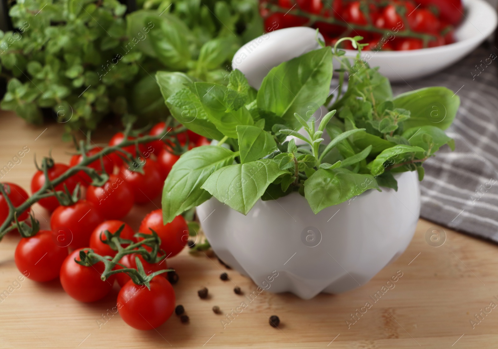 Photo of Mortar with fresh herbs, cherry tomatoes and black peppercorns on wooden table, closeup