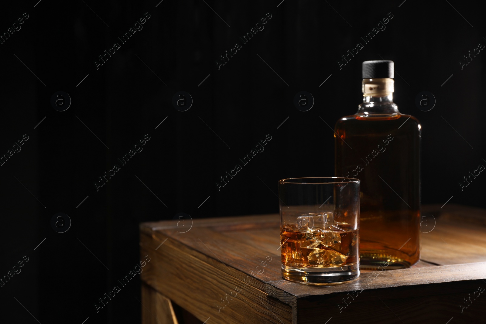 Photo of Whiskey with ice cubes in glass and bottle on wooden crate against black background, space for text