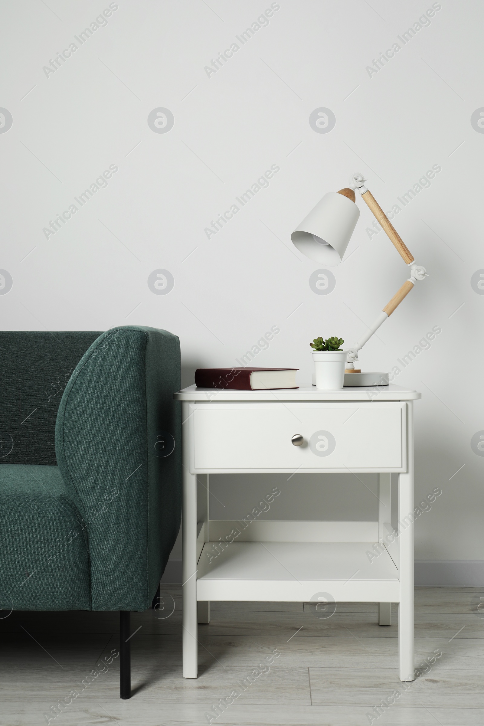 Photo of Stylish lamp, houseplant and book on white side table near soft sofa in light room. Interior design