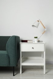 Stylish lamp, houseplant and book on white side table near soft sofa in light room. Interior design